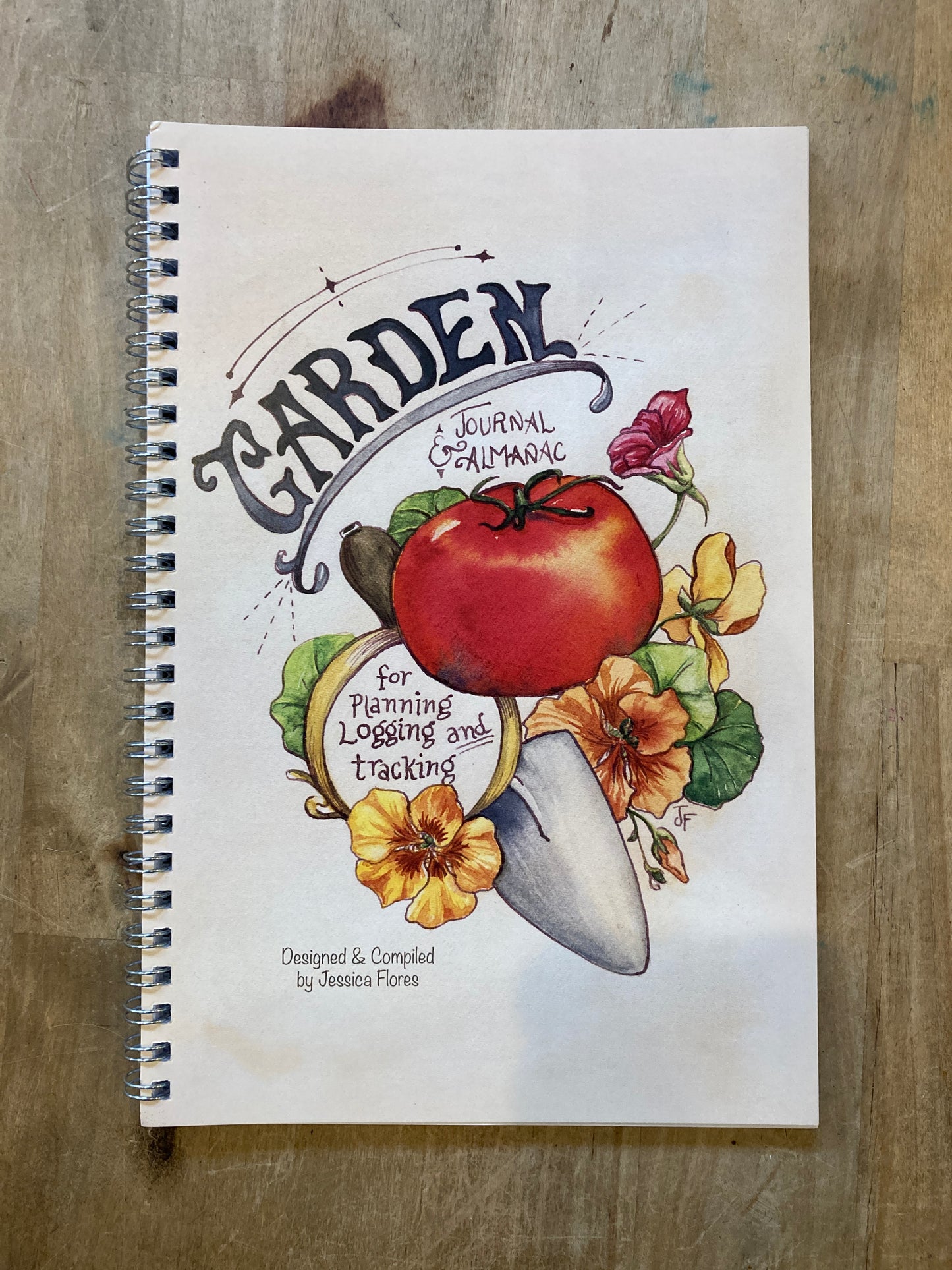 White wire-bound notebook. Cover illustration of a tomato, flowers, and trowel. Text reads "Garden journal & almanac for planning, logging, and tracking."